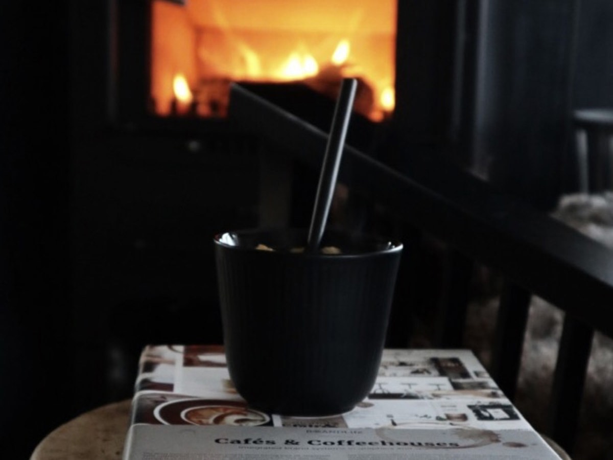 En kopp med varm kakao står på et mørkt bord foran en peis med flammer